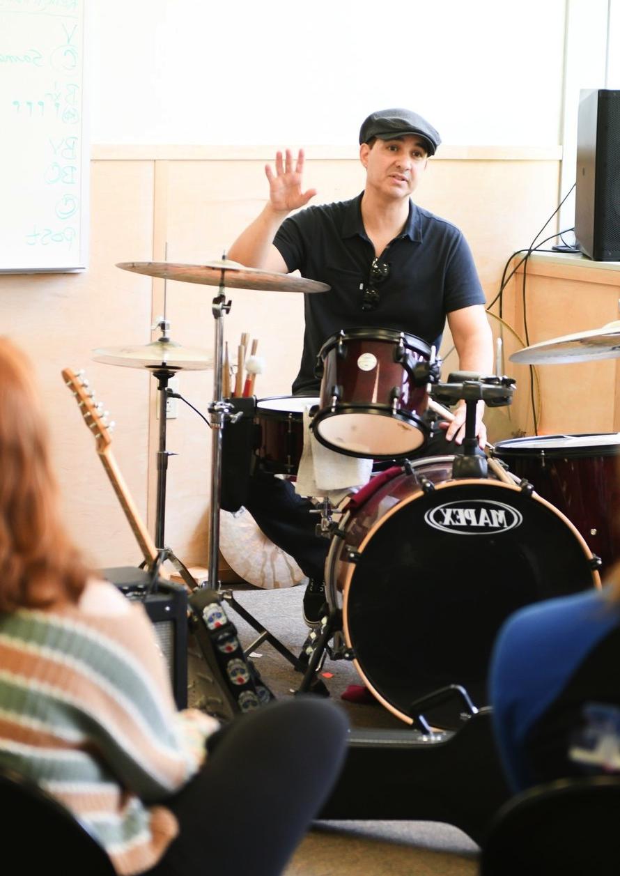 A music instructor wearing a black shirt and a cap is sitting at a drum set, raising his hand mid-explanation to a student. The setting appears to be an informal music class with a whiteboard in the background, enhancing the educational atmosphere.