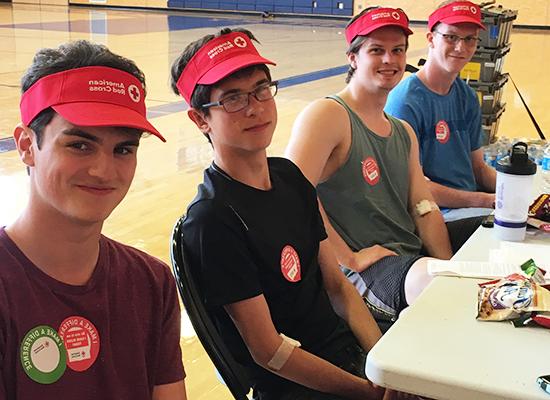 Four young men with light skin, wearing red American Red Cross visors, sitting at a table in a gymnasium. They are wearing casual clothing and have stickers indicating they participated in a blood donation event.